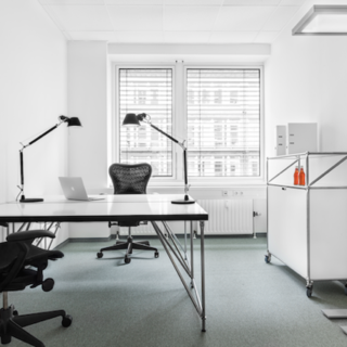 Ein moderner, minimalistischer Büroraum mit einem Schreibtisch, einem schwarzen Bürostuhl und zwei Tischlampen. Auf dem Schreibtisch steht ein Laptop. In der Ecke befindet sich ein rollbarer, weißer Schrank mit zwei Getränkeflaschen. Große Fenster sorgen für Tageslicht.