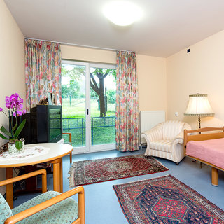 Ein helles Zimmer mit einem Einzelbett, einem bequemen Sessel und einem Tisch. An einem Fenster hängen bunte Vorhänge. Auf dem Tisch steht eine Orchidee, und ein Teppich liegt auf dem Boden. Draußen ist eine grüne Wiese mit Bäumen zu sehen.