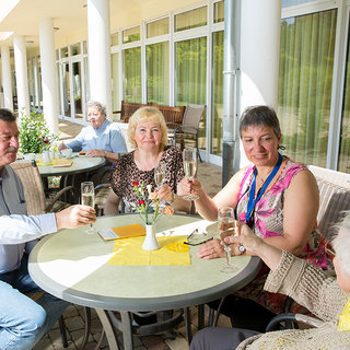 Eine Gruppe von fünf Personen sitzt an einem Tisch im Freien und stößt mit Gläsern an. Die Atmosphäre ist freundlich und feierlich. Im Hintergrund sind weitere Personen zu sehen, die sich unterhalten. Es ist tagsüber und die Umgebung wirkt einladend und hell.