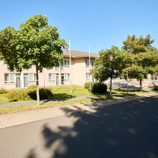 Ein helles Gebäude mit mehreren Fenstern befindet sich neben einer gepflasterten Straße. Der Garten ist grün und von Bäumen umgeben. Der Himmel ist klar und blau.
