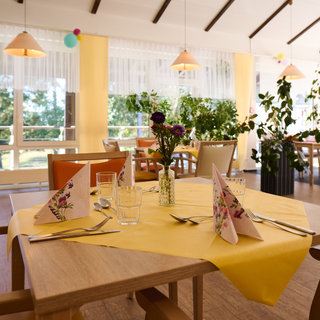 Ein helles Restaurant mit einer einladenden Atmosphäre. Auf einem Holzttisch liegt eine gelbe Tischdecke, geschmückt mit Servietten und einem kleinen Blumenstrauß. Das Restaurant ist lichtdurchflutet, umgeben von Pflanzen und großen Fenstern, die einen Blick nach draußen ermöglichen.