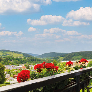 Balkon mit blühenden roten Geranien, Ausblick auf eine grüne Hügellandschaft unter einem blauen Himmel mit weißen Wolken. Im Hintergrund sind Dörfer und sanfte Hügel sichtbar.