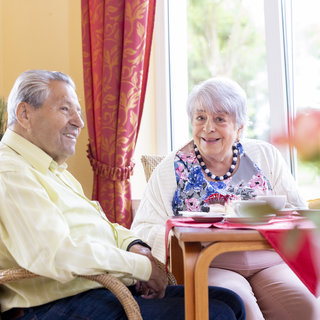 Ein älteres Paar sitzt lächelnd an einem Tisch in einem hellen Raum. Sie genießen gemeinsam eine Tasse Kaffee oder Tee. Die Atmosphäre ist freundlich und einladend, mit Blick auf einen Garten durch ein Fenster.