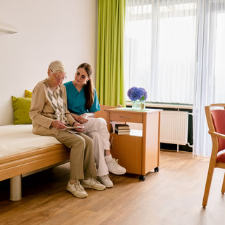 Eine ältere Frau sitzt auf einem Bett und betrachtet gemeinsam mit einer Pflegekraft Dokumente. Die Atmosphäre ist freundlich und unterstützend, mit heller Beleuchtung und einem gemütlichen Raumdesign. Im Hintergrund sind ein Tisch und eine Vase mit Blumen zu sehen.