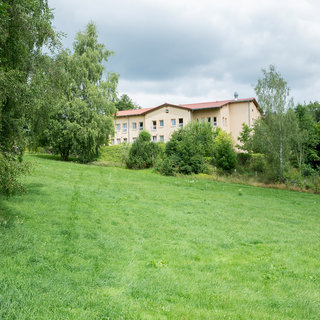 Ein mehrstöckiges Gebäude steht auf einer Wiese, umgeben von Bäumen und Grünflächen. Der Himmel ist bewölkt, und die Landschaft wirkt ruhig und naturnah.