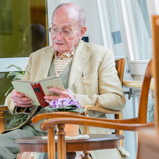 Ein älterer Mann sitzt in einem bequemen Stuhl und liest ein Buch. Er trägt eine beige Jacke und eine karierte Bluse. Auf einem kleinen Tisch neben ihm steht ein Blumentopf mit einer blühenden Pflanze. Der Raum wirkt hell und einladend.