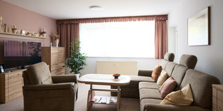 Das Bild zeigt ein helles Wohnzimmer mit zwei bequemen Sofas, einem Couchtisch und einer Pflanze. Ein Fernseher steht auf einem Holzschrank, und ein großes Fenster mit Vorhängen lässt viel Tageslicht herein. Die Wände sind in sanften Farben gestaltet.