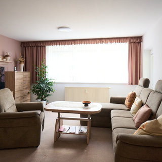 Das Bild zeigt ein helles Wohnzimmer mit zwei bequemen Sofas, einem Couchtisch und einer Pflanze. Ein Fernseher steht auf einem Holzschrank, und ein großes Fenster mit Vorhängen lässt viel Tageslicht herein. Die Wände sind in sanften Farben gestaltet.