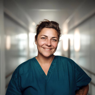 Eine lächelnde Frau in einem grünen Krankenhauskittel steht in einem gut beleuchteten Korridor. Der Hintergrund ist verschwommen, was den Fokus auf ihr freundliches Gesicht lenkt.