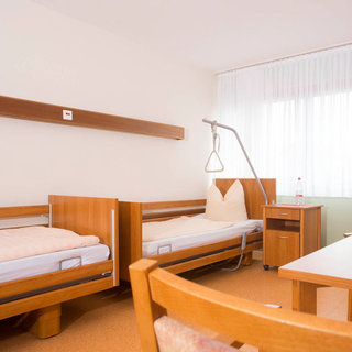 Das Bild zeigt ein schlicht eingerichtetes Krankenhauszimmer mit zwei Betten, einem kleinen Tisch und einem Stuhl. Die Wände sind hell gestrichen, und große Fenster sorgen für Tageslicht. Es gibt eine Halterung über einem der Betten, vermutlich für medizinische Zwecke.