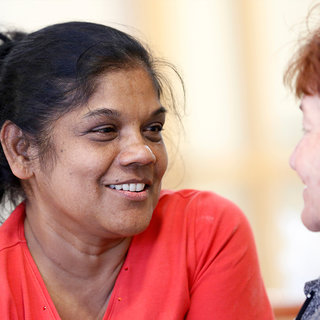 Zwei Frauen in einem freundlichen Gespräch. Eine Frau mit dunklem Haar und rotem Oberteil lächelt und schaut die andere Frau an, die eine rötliche Frisur hat. Beide strahlen eine positive und einladende Atmosphäre aus.