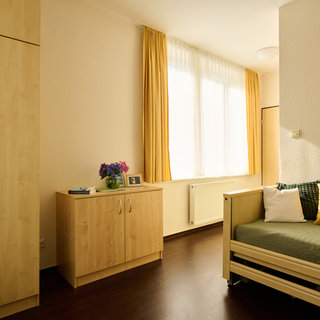 Ein helles Zimmer mit einem Bett, das mit grüner Bettwäsche und bunten Kissen dekoriert ist. An der Wand steht ein Holzschrank, neben einem kleinen Tisch mit einer Blumenvase und einem Bilderrahmen. Das Fenster lässt viel Licht herein, und die Vorhänge sind gelb.