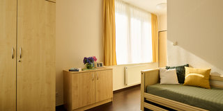 Ein helles Zimmer mit einem Bett, das mit grüner Bettwäsche und bunten Kissen dekoriert ist. An der Wand steht ein Holzschrank, neben einem kleinen Tisch mit einer Blumenvase und einem Bilderrahmen. Das Fenster lässt viel Licht herein, und die Vorhänge sind gelb.