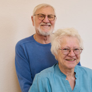 Ein älteres Paar lächelt freundlich in die Kamera. Der Mann steht hinter der Frau, beide tragen Brillen und haben graue Haare. Die Frau hat eine helle Bluse an, während der Mann einen blauen Pullover trägt. Der Hintergrund ist schlicht und hell.