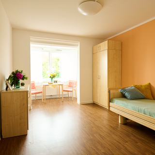Ein helles Zimmer mit einem Einzelbett, einem Holzschrank und einem Tisch mit zwei Stühlen. Die Wände sind teilweise orange gestrichen, und ein Fenster lässt Tageslicht herein. Auf dem Tisch steht eine Vase mit Blumen.