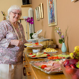 Eine fröhliche ältere Frau steht an einem Tisch mit verschiedenen Platten voller frischer Früchte und Häppchen. Sie lächelt, während sie eine Servierplatte mit leckeren Snacks in der Hand hält. Im Hintergrund sind Blumen und eine dekorative Vase sichtbar.