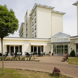 Eine moderne Gebäudeansicht mit einem Gartenbereich. Es gibt Tische und Stühle im Freien sowie eine gepflasterte Gehweg. Im Hintergrund sind hohe Wohngebäude sichtbar. Der Eingang hat große Glasflächen und ist von Pflanzen umgeben.