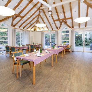 Ein hell erleuchteter Speisesaal mit Holzbalkendecke. Mehrere Tische sind deckend mit lila Tischdecken und Geschirr eingedeckt. Fenster bieten einen Blick nach draußen in einen grünen Garten. Der Raum vermittelt eine einladende und gemütliche Atmosphäre.