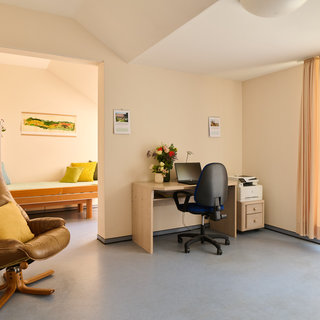 Ein helles Zimmer mit einem Arbeitsbereich. Ein Schreibtisch mit einem Stuhl und einem Computer steht neben einem Drucker. In der Ecke befindet sich ein bequemer Sessel mit einem gelben Kissen. Im Hintergrund ist ein Bett sichtbar und Fenster mit Vorhängen lassen Tageslicht herein.