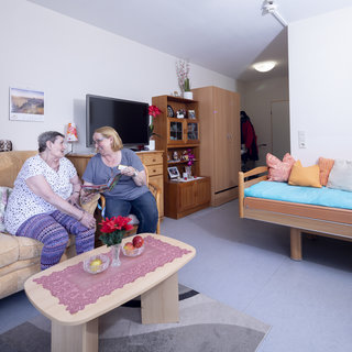 Zwei Frauen sitzen auf einer Sofa in einem gemütlichen Wohnraum. Sie unterhalten sich freundlich und teilen einen Moment des Lachens. Ein kleiner Tisch mit frischen Blumen und Snacks ergänzt die entspannte Atmosphäre des Zimmers.