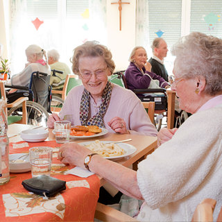 Zwei ältere Frauen sitzen an einem gedeckten Tisch, lächeln und essen zusammen. Im Hintergrund sind weitere Bewohner eines Altenheims zu sehen, die in einer hellen, einladenden Umgebung speisen. Die Atmosphäre ist freundlich und gesellig.