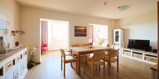 Ein heller Wohnraum mit einem großen Holztisch und sechs Stühlen. An einer Wand steht ein Fernseher und ein Sideboard. Durch die Fenster fällt Tageslicht, und einige Pflanzen sind im Raum verteilt. An einer Wand hängt ein Bild von einer blühenden Landschaft.