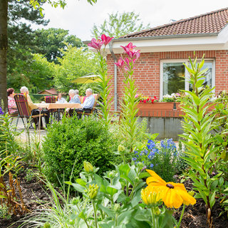 Eine Gruppe von fünf Personen sitzt an einem Tisch im Freien, umgeben von blühenden Pflanzen und Sträuchern. Im Hintergrund ist ein helles Haus mit großen Fenstern zu sehen. Die Szene vermittelt eine entspannte, sommerliche Atmosphäre.