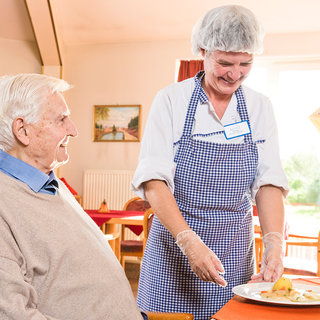 Eine lächelnde Servicemitarbeiterin in schützender Kleidung reicht einem älteren Mann einen Teller mit Essen. Der Mann sitzt auf einem Stuhl und schaut freundlich zur Mitarbeiterin. Die Umgebung wirkt hell und einladend, möglicherweise in einem Speisesaal oder einer Einrichtung für Senioren.