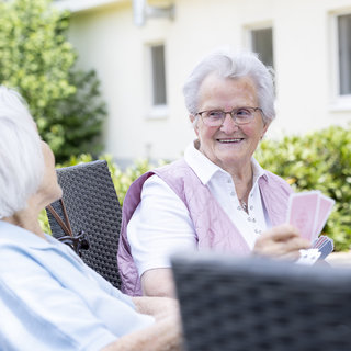 Zwei ältere Frauen sitzen draußen auf einem Balkon. Die Frau auf der rechten Seite lächelt und hält einige Karten in der Hand, während die andere Frau sie ansieht. Im Hintergrund sind grüne Pflanzen und ein Gebäude sichtbar.
