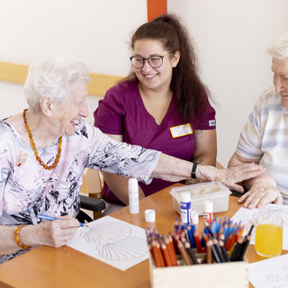 Zwei ältere Damen sitzen an einem Tisch und malen mit Buntstiften. Eine Pflegekraft lächelt und unterstützt sie. Auf dem Tisch liegen Malvorlagen und Farben. Die Atmosphäre ist freundlich und einladend, was auf kreative Aktivitäten im Seniorenheim hindeutet.