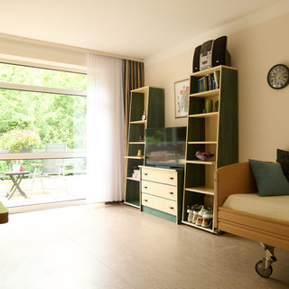 Ein helles Zimmer mit einem Pflegebett, einem Fernsehen und einem Bücherregal. Rechts steht ein Tisch mit einer blauen Dekoration. Große Fenster lassen Tageslicht herein und bieten einen Blick auf einen begrünten Außenbereich.