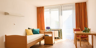 Ein heller Raum mit Holzboden, ausgestattet mit einem Einzelbett, einem kleinen Tisch und einem Stuhl. Vor den Fenstern hängen orangerote Vorhänge. Auf dem Tisch stehen Pflanzen und ein Blumentopf. Das Licht fällt durch das Fenster und sorgt für eine einladende Atmosphäre.