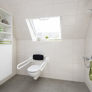 Ein modernes Badezimmer mit einem barrierefreien WC. Es ist hell gestaltet mit großen Fliesen und einem Fenster. Neben dem WC befindet sich ein Haltegriff, sowie ein Handtuchhalter mit einem grünen Handtuch. Ein Regal mit gefalteten Handtüchern vervollständigt den Raum.
