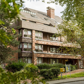 Ein mehrstöckiges Wohngebäude aus roten Ziegeln mit großen Fenstern. Die Fassade ist mit Schiefermaterial verkleidet, und es gibt einen eingezäunten Gartenbereich mit Büschen. Ein Briefkasten ist an der Vorderseite sichtbar.