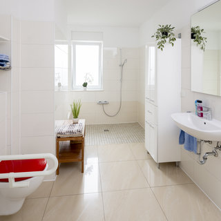Ein modernes Badezimmer mit hellen Fliesen. Auf der linken Seite befindet sich eine barrierefreie Toilette mit Haltegriffen. Neben dem Fenster ist eine Dusche mit einer praktischen Duschstange. Ein weißes Waschbecken mit Spiegel und ein Regal mit Handtüchern sind ebenfalls vorhanden. Grüne Pflanzen sorgen für eine freundliche Atmosphäre.