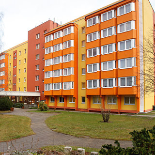 Ein mehrstöckiges Gebäude in Gelb- und Orangetönen. Im Vordergrund befindet sich ein grüner Garten mit einem Weg, der zur Eingangstür führt. Links steht ein roter Feuerhydrant, umgeben von niedrigem Bewuchs. Die Fenster des Gebäudes sind klar und einladend.