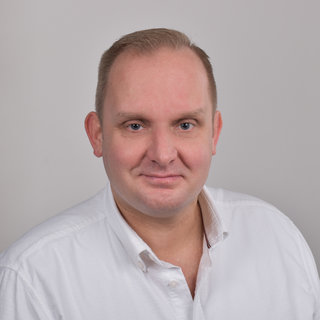 Ein Smiling Mann mit kurzen, hellen Haaren trägt ein weißes Hemd. Er schaut direkt in die Kamera vor einem neutralen Hintergrund. Sein Gesichtsausdruck vermittelt Freundlichkeit und Offenheit.