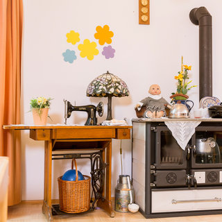 In einer traditionellen Küche steht ein Tisch mit einer Nähmaschine und einer dekorativen Lampe. Neben dem Tisch befindet sich ein Herd mit einem Tischgeschirr, einer Puppe und einer Vase mit Blumen. An der Wand sind bunte Blütenmuster angebracht.