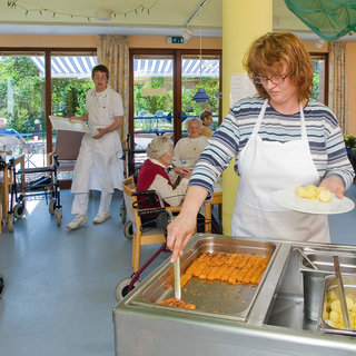 Eine Mitarbeiterin in einer Einrichtung für ältere Menschen serviert Essen aus einer Buffet-Wanne, während ein Kollege im Hintergrund Geschirr trägt. Im Raum arbeiten weitere Personen, und einige ältere Menschen sitzen in Stühlen oder Rollstühlen. Fenster lassen natürliches Licht herein.