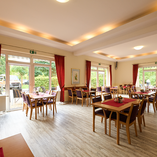 Der Innenraum eines Restaurants mit hellen Wänden, Holzfußboden und großen Fenstern, die einen Blick ins Freie ermöglichen. Tische mit roten Tischtüchern und Stühlen sind gleichmäßig im Raum angeordnet. Vorhänge in Rot rahmen die Fenster ein.
