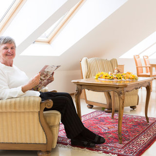 Eine lächelnde ältere Frau sitzt in einem bequemen Sessel und hält eine Zeitschrift in der Hand. Um sie herum sind ein Tisch mit einem Blumengesteck und ein Teppich. Die Wände sind hell und es gibt ein Fenster, das viel Licht hereinlässt.