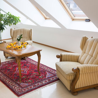 Ein gemütlicher Raum mit zwei gestreiften Sesseln und einem Holztisch in der Mitte. Auf dem Tisch steht eine Vase mit Sonnenblumen. Der Boden ist mit einem roten Teppich ausgelegt und Fenster mit schrägen Dächern lassen Tageslicht herein.