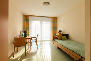 Ein einfaches Zimmer mit einem Einzelbett, einem Tisch und einem Stuhl. Vor den großen Fenstern hängen bunte Vorhänge. Auf dem Tisch steht eine Vase mit Blumen. Der Boden ist hell und der Raum wirkt freundlich und einladend.