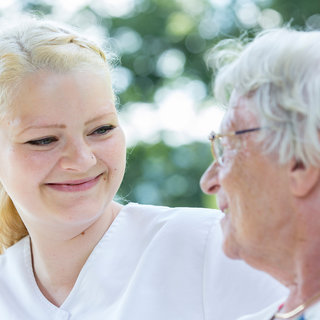 Eine junge Frau und eine ältere Dame sitzen sich gegenüber und lächeln sich freundlich an. Die Atmosphäre ist warm und herzlicher Austausch zu spüren. Die Umgebung ist hell und grünes Laub ist im Hintergrund zu erkennen.