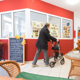 Eine Person mit Gehhilfe bewegt sich durch einen hellen Raum mit roter Wand, umgeben von Tisch und Stühlen. An den Wänden hängen Bilder, und ein schwarzes Schild informiert über eine Veranstaltung. Der Raum wirkt einladend und freundlich.