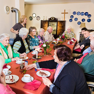 Eine Gruppe älterer Menschen sitzt an einem langen Tisch, gedeckt mit Kuchen und Getränken. Sie haben freundliche Gesichter und unterhalten sich. Eine Frau steht am Kopf des Tisches und lächelt. Blumen dekorieren den Tisch. Die Atmosphäre ist fröhlich und gesellig.
