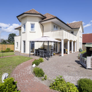 Eine helle, moderne Villa mit einem begrünten Garten und einem gepflasterten Weg. Vor dem Haus stehen ein Unterstand mit Tisch und Stühlen sowie ein Strandkorb. Im Hintergrund ist ein blauer Himmel mit einigen Wolken sichtbar.