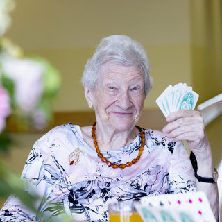 Eine ältere Dame sitzt an einem Tisch und hält lächelnd eine Hand voller Spielkarten. Sie trägt ein farbenfrohes Oberteil und eine Halskette. Im Hintergrund sind verschwommene Blumen und eine gemütliche Raumgestaltung sichtbar.