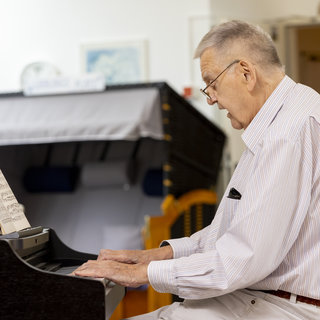 Ein älterer Mann sitzt am Klavier und spielt, während er Notenblätter betrachtet. Er trägt eine Brille und ein Hemd mit langen Ärmeln. Der Raum scheint hell und freundlich zu sein.