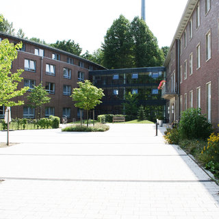 Ein Innenhof mit zwei Gebäuden, eines aus roten Ziegelsteinen und das andere modern mit großen Fenstern. In der Mitte stehen einige Bäume und Sträucher, umgeben von Pflastersteinen. Der Bereich ist hell erleuchtet, mit viel Tageslicht und grüner Vegetation.